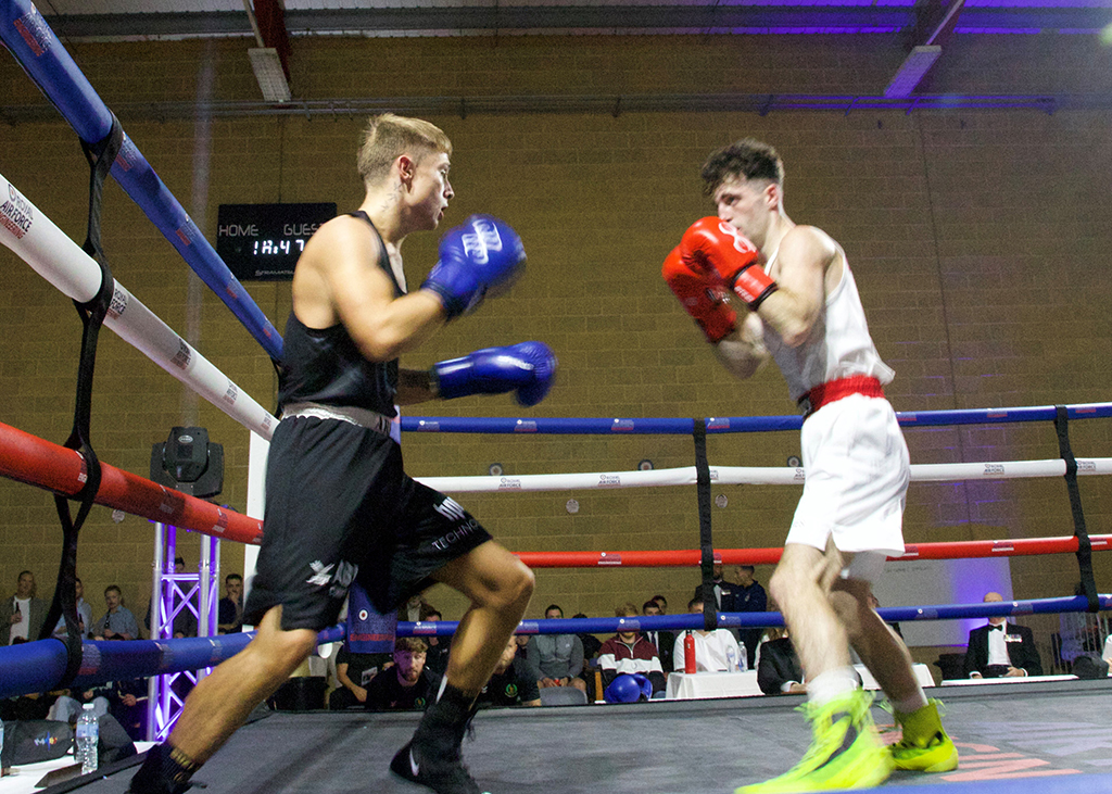 On the left, an Army boxer wearing black is in motion. On the right, a Royal Navy boxer wearing white with a red waistband stands with boxing gloves raised defensively. The match takes place in a ring with red, white, and blue ropes, surrounded by a seated audience and event staff in the background under indoor arena lighting.